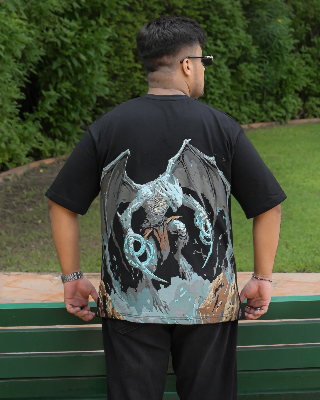 A man viewed from behind wearing a black t-shirt with a large graphic print of a winged creature holding a glowing blue orb, standing outdoors in front of a green bench and foliage.