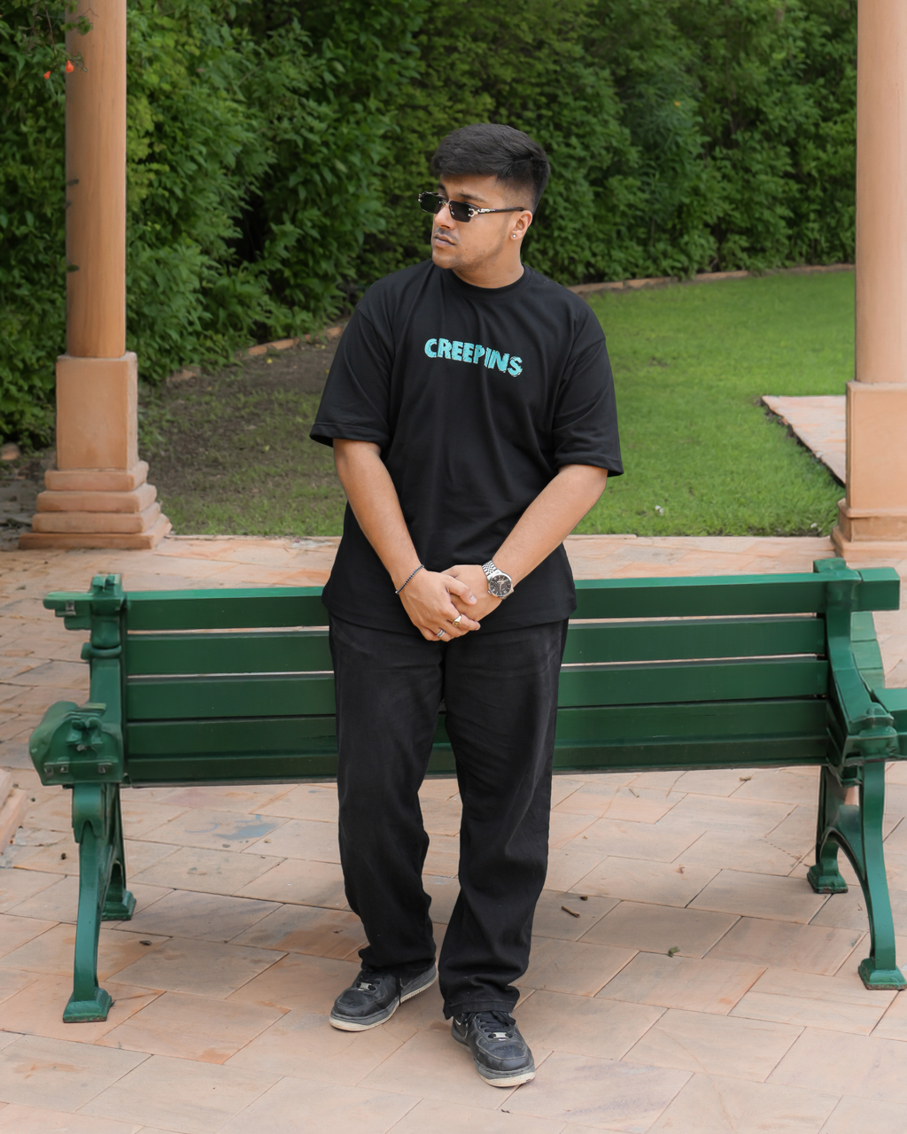 A man wearing a black t-shirt with 'CREEPINS' printed in light blue on the chest, standing on a brick patio in front of a green park bench and lush green trees.