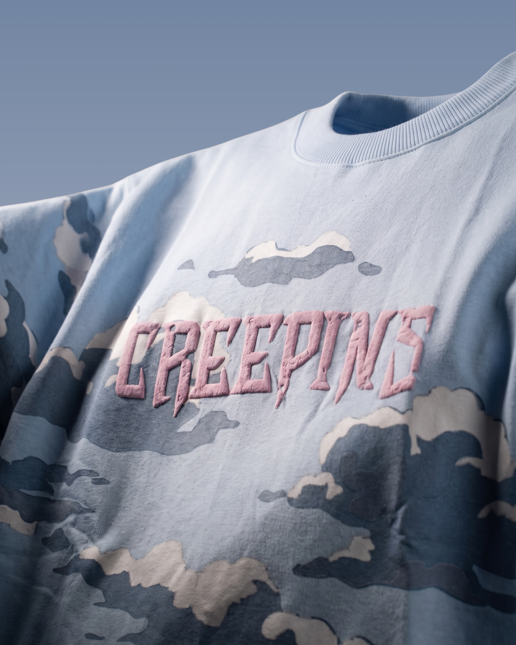 A close-up of a light blue crewneck sweatshirt featuring a blue, gray, and beige camouflage pattern and the word 'CREEPINS' embroidered across the chest in a pink gothic font.