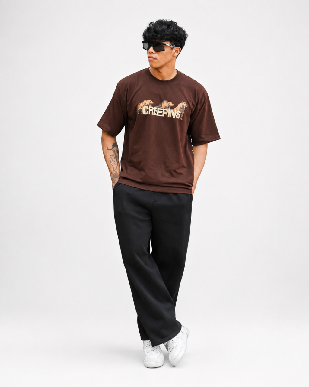 A man modeling a brown crewneck t-shirt with a 'CREEPINS' graphic across the chest, paired with black sweatpants and white sneakers, set against a plain light gray studio background.