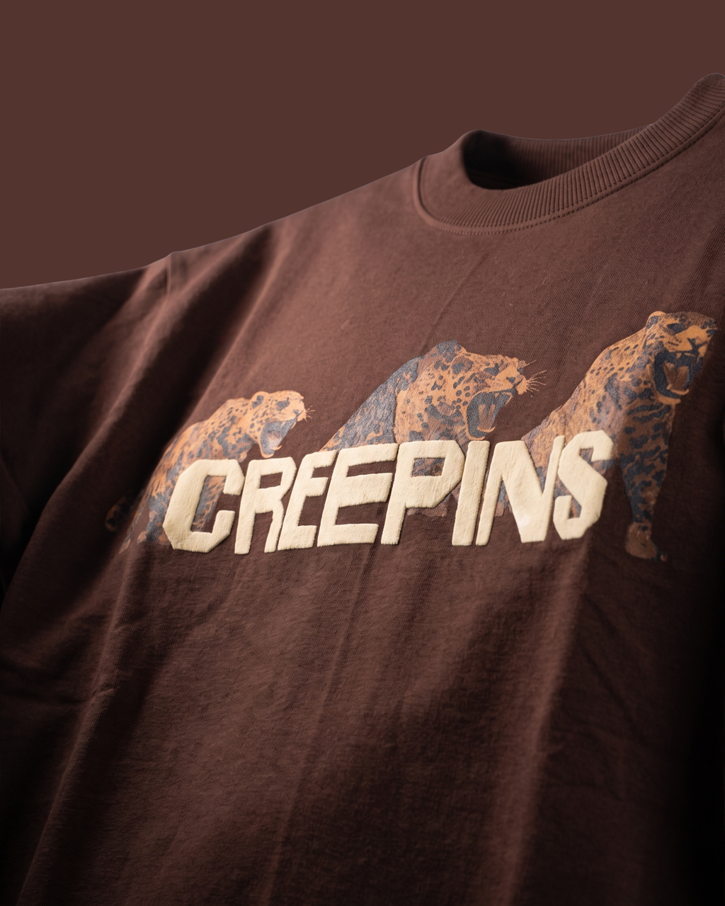 A close-up of a dark brown crewneck sweatshirt featuring a graphic print of three bears in a distressed tan and blue pattern, with the word 'CREEPINS' printed in large cream-colored letters across the chest.
