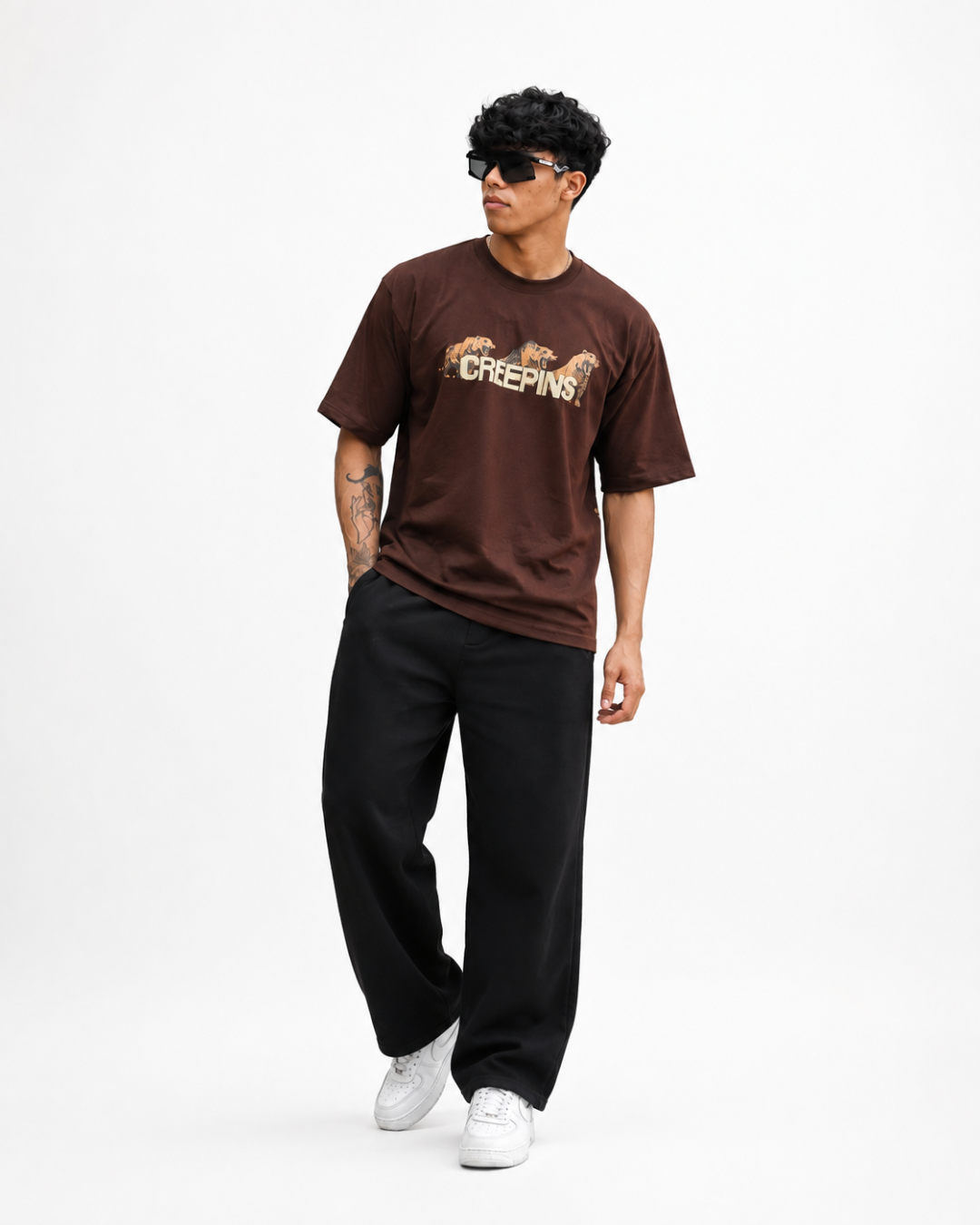 A man modeling a brown short-sleeve graphic t-shirt with 'CREEPINS' and horse artwork across the chest, paired with black sweatpants and white sneakers, against a plain white studio background.
