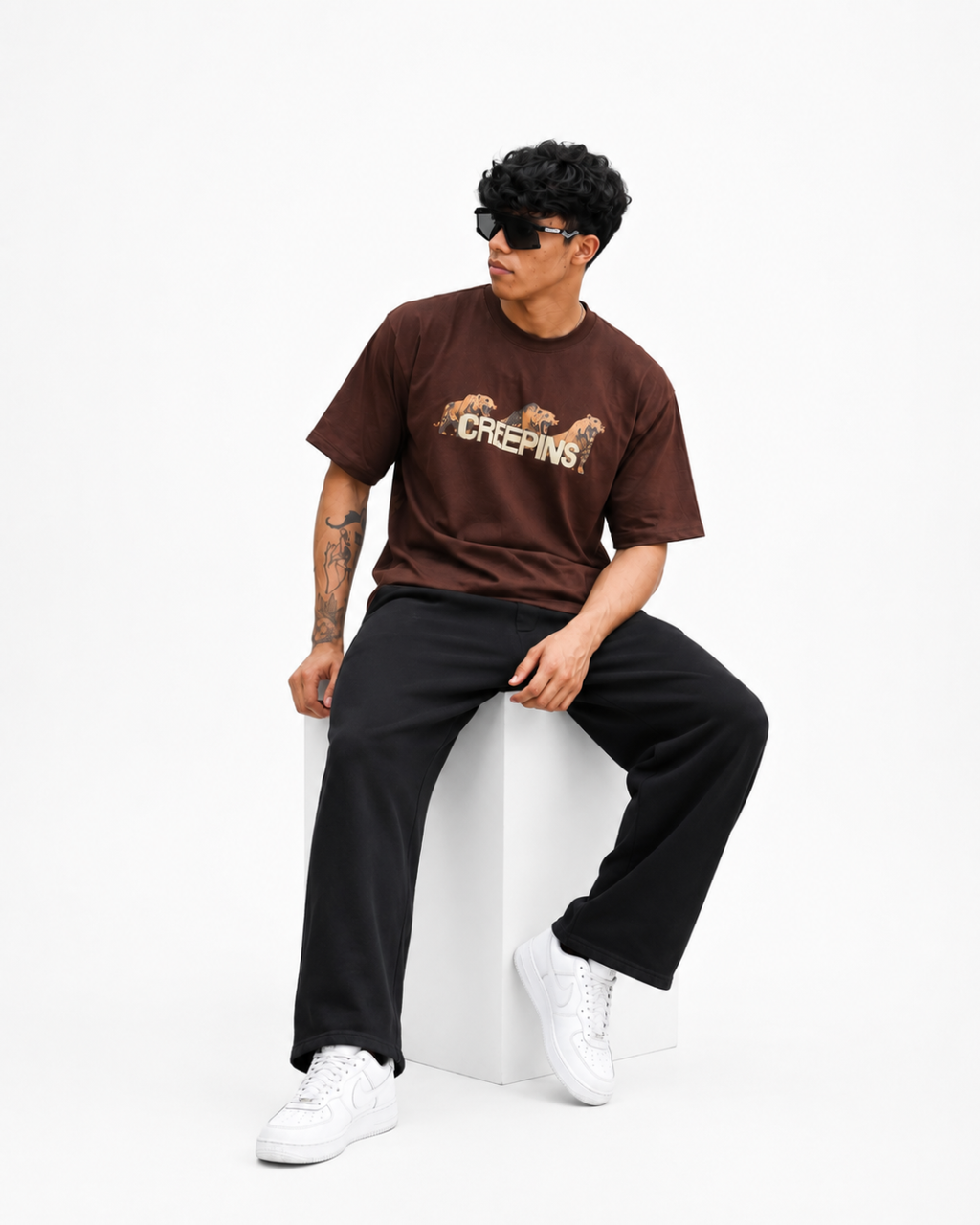 A man wearing a brown Creepins t-shirt, black sweatpants, and white sneakers sits on a white block against a white studio background.