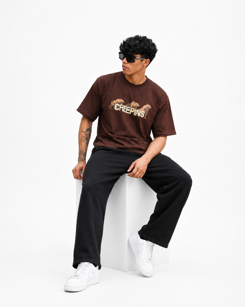 A man wearing a brown Creepins t-shirt, black sweatpants, and white sneakers sits on a white block against a white studio background.