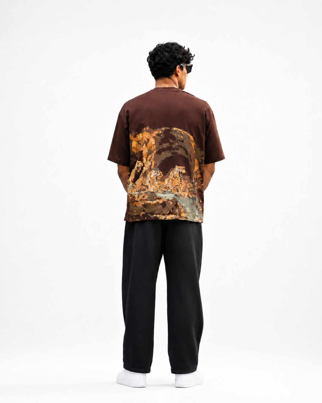 A man viewed from behind wearing a brown short-sleeve t-shirt with a large, detailed landscape-style graphic print in orange, gold, and blue tones across the back, paired with black pants and white socks, against a plain white studio background.