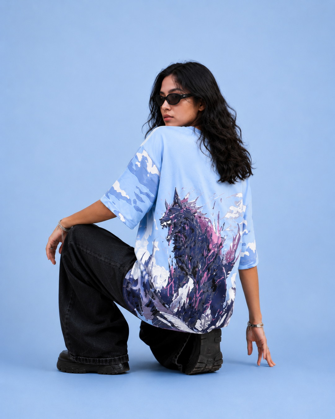 A model wearing an oversized light blue t-shirt with a large purple and black monster graphic on the back, posing on a solid blue studio background.