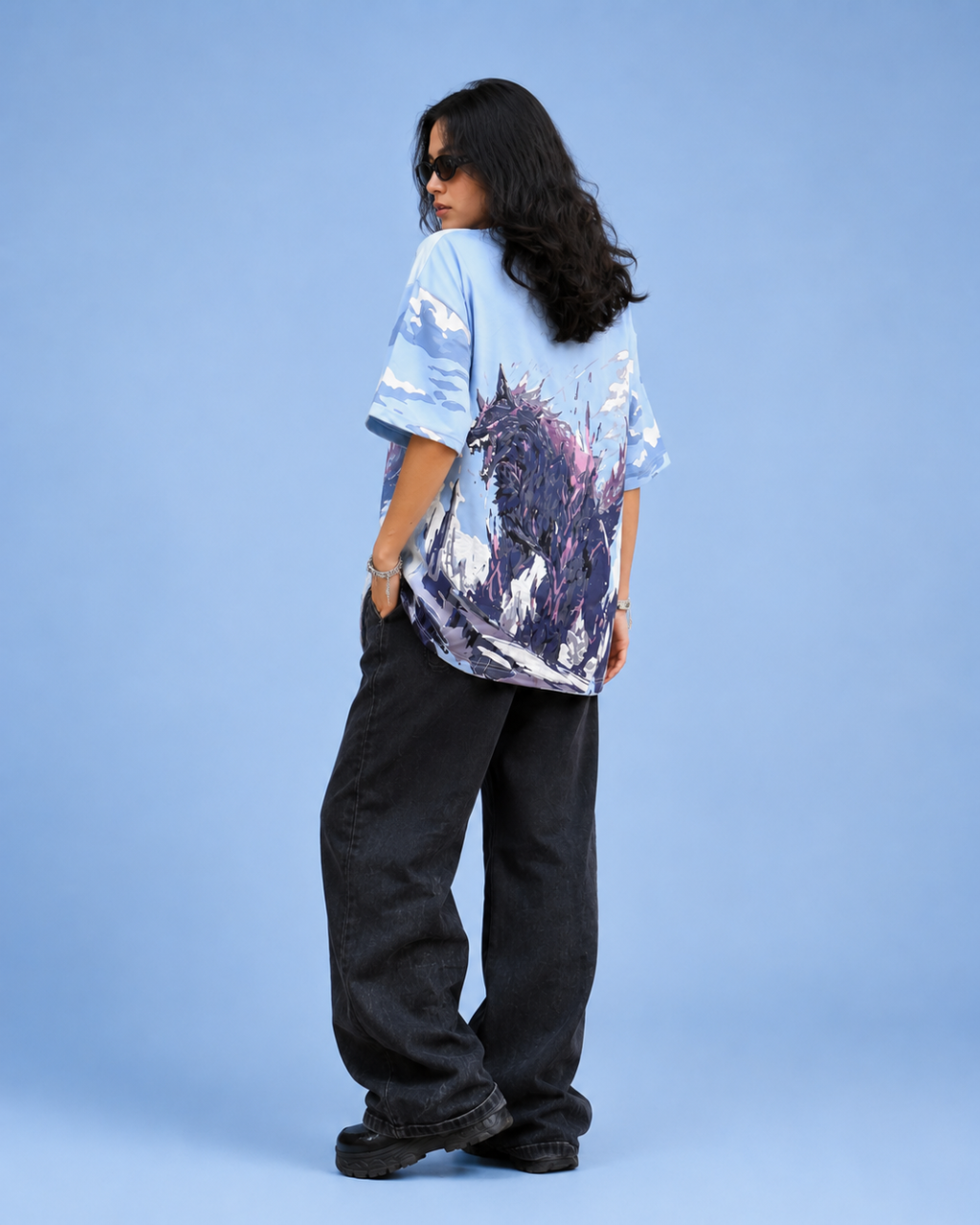 A model is shown from the back wearing a light blue oversized t-shirt with a dark purple and black graphic print, paired with loose black pants, against a solid light blue background.