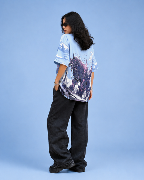 A model is shown from the back wearing a light blue oversized t-shirt with a dark purple and black graphic print, paired with loose black pants, against a solid light blue background.