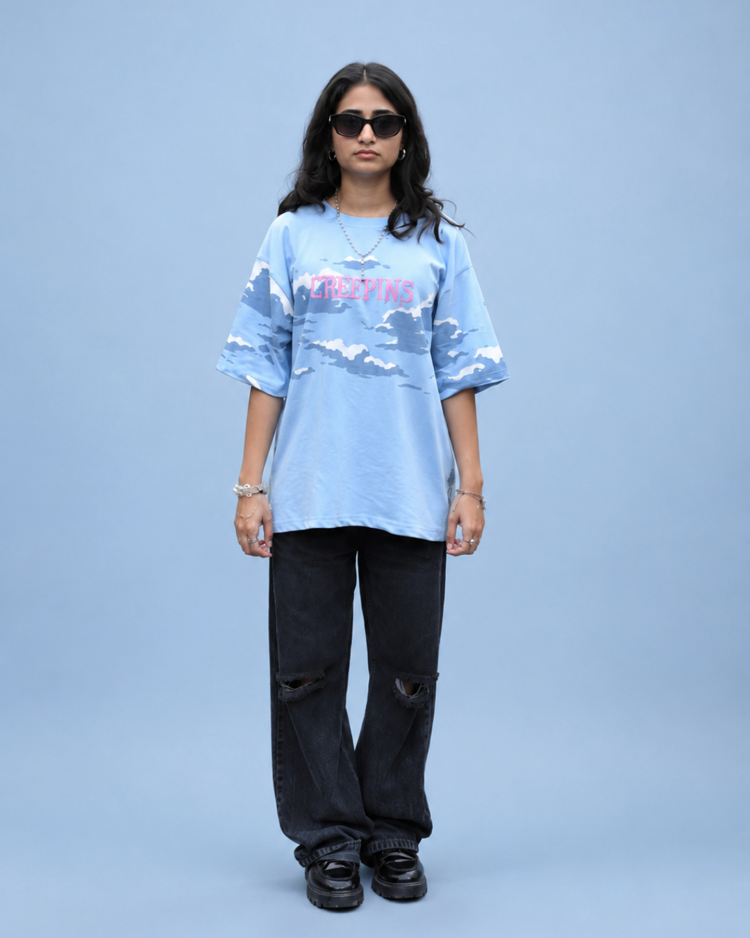 A model stands against a solid light blue studio background wearing a light blue graphic t-shirt with a pink and white text design, black distressed jeans, and black sneakers.