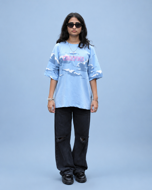 A model stands against a solid light blue studio background wearing a light blue graphic t-shirt with a pink and white text design, black distressed jeans, and black sneakers.